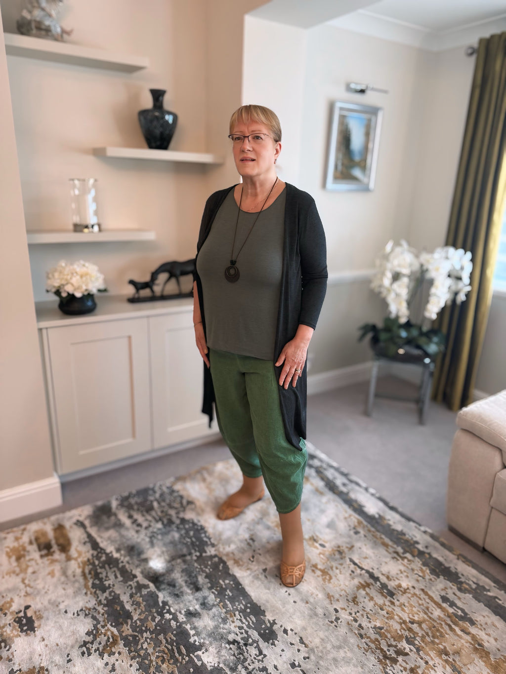 Woman wearing a longline Italian made cardigan and harem pants with vest standing in a living room with decor elements like shelves, vases, and a rug.