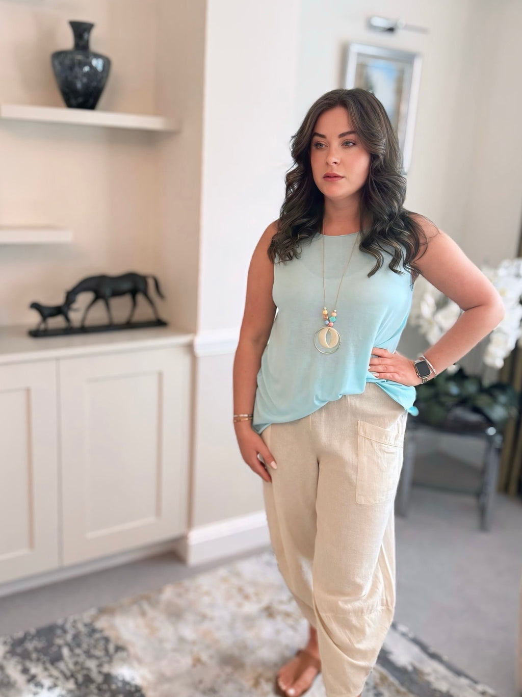 Woman standing in a living room wearing a light blue sleeveless top and cotton harem beige pants.
