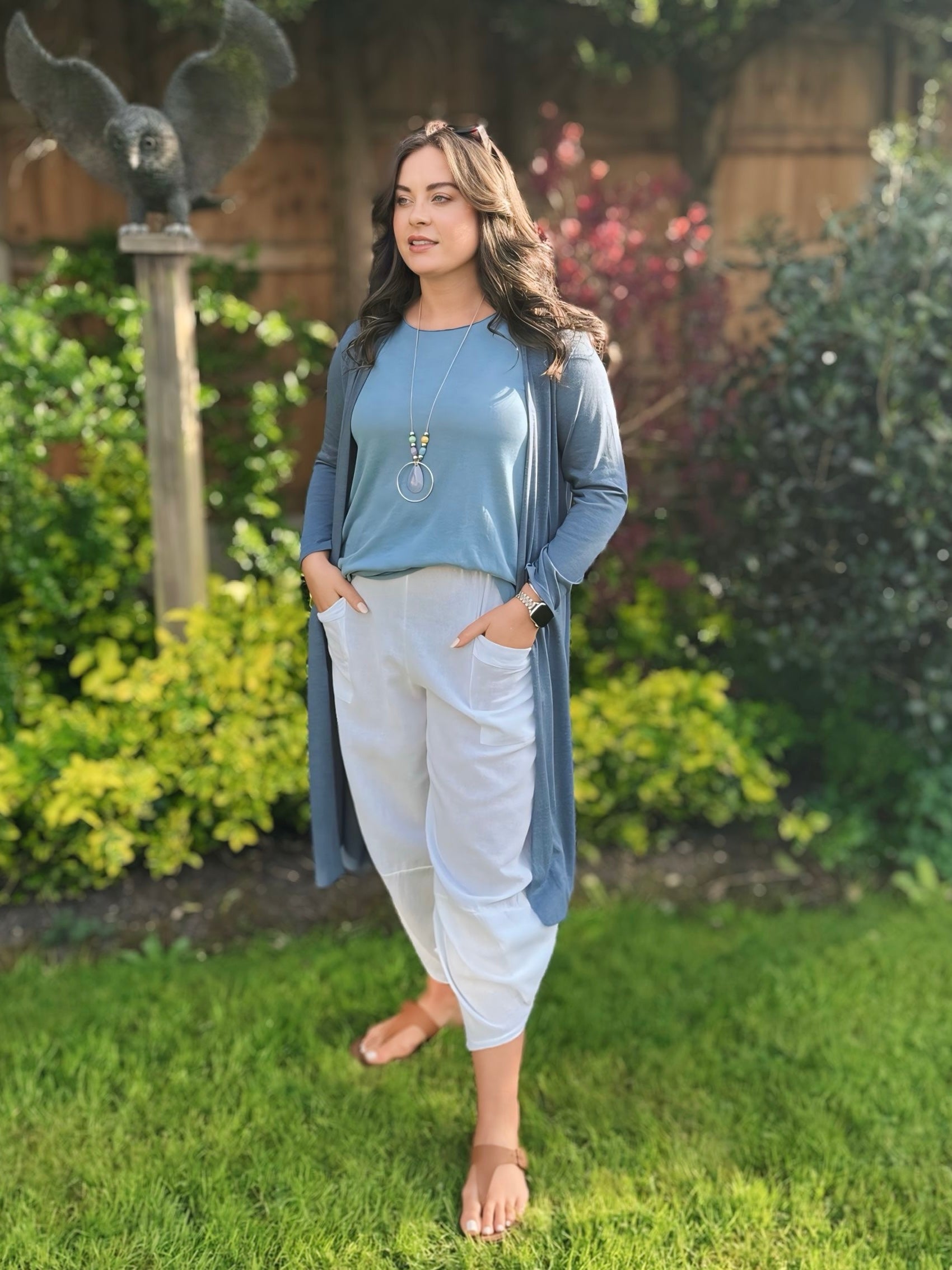 Woman in a blue long line cardigan and white cotton harem pants standing in a garden.