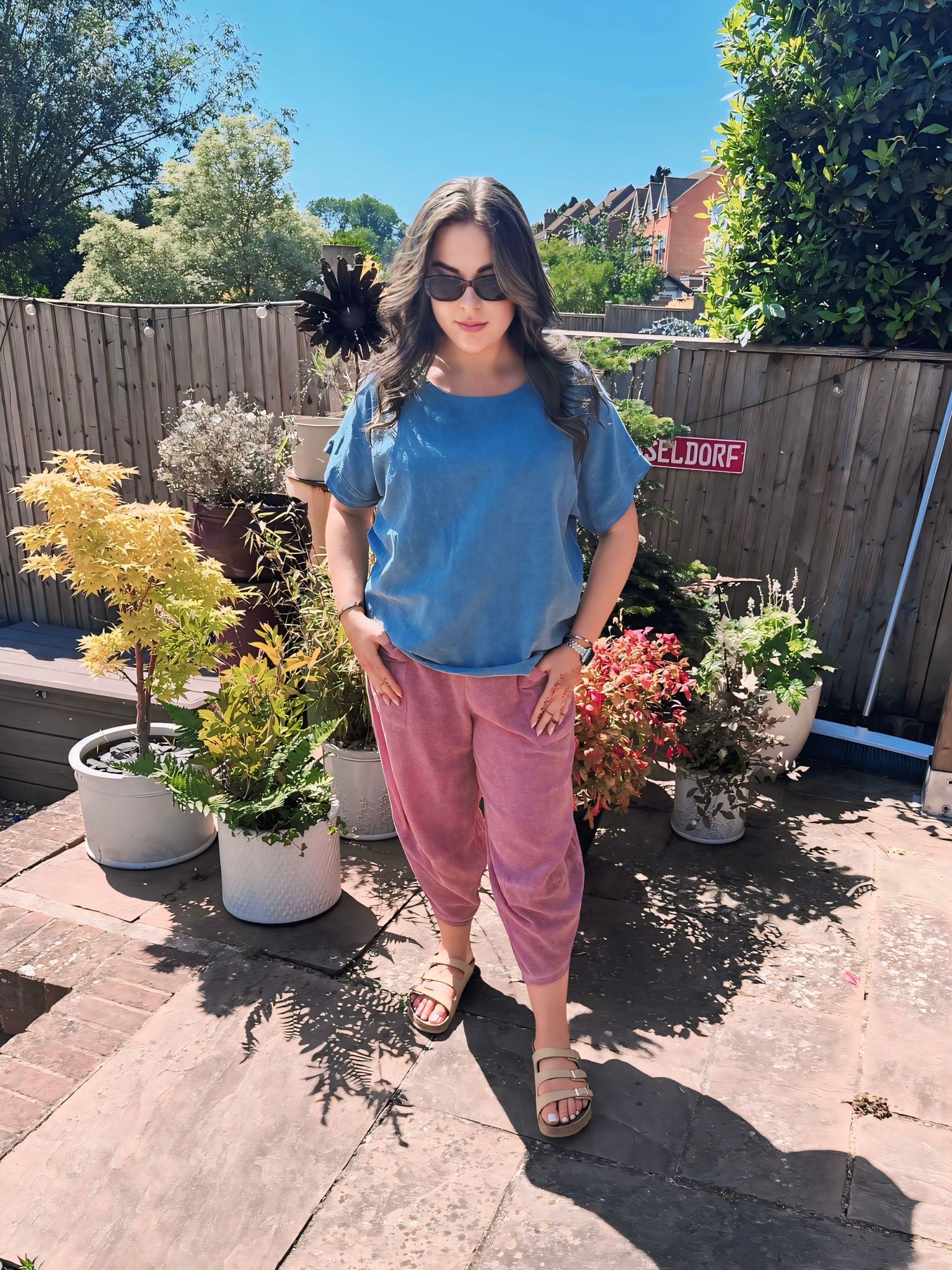 Woman standing in a garden with potted plants on a sunny day wearing a blue cotton top and dusky pink cotton harem pants